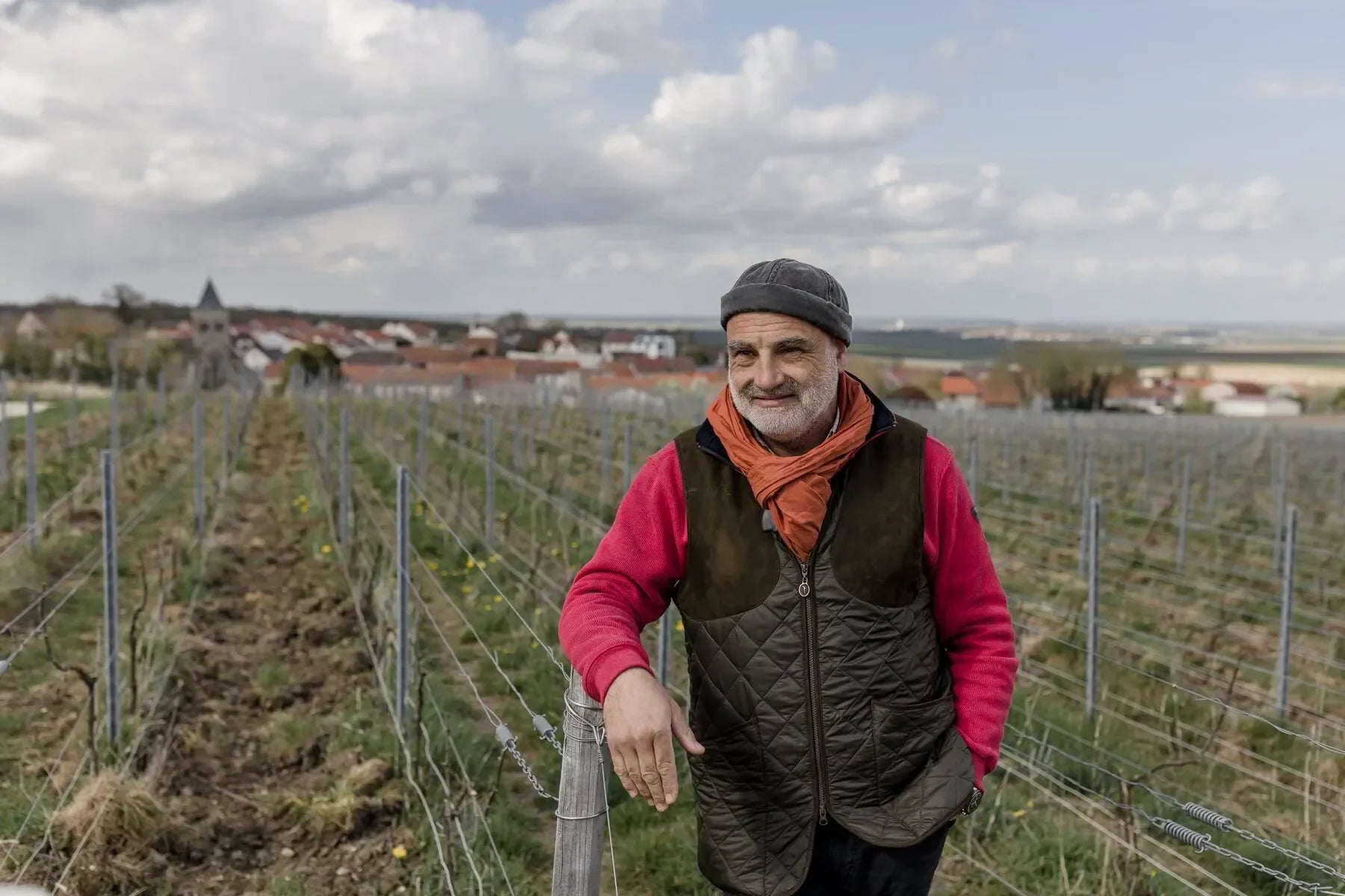 Man in vineyard wearing red jacket and orange scarf, with village and cloudy sky in background