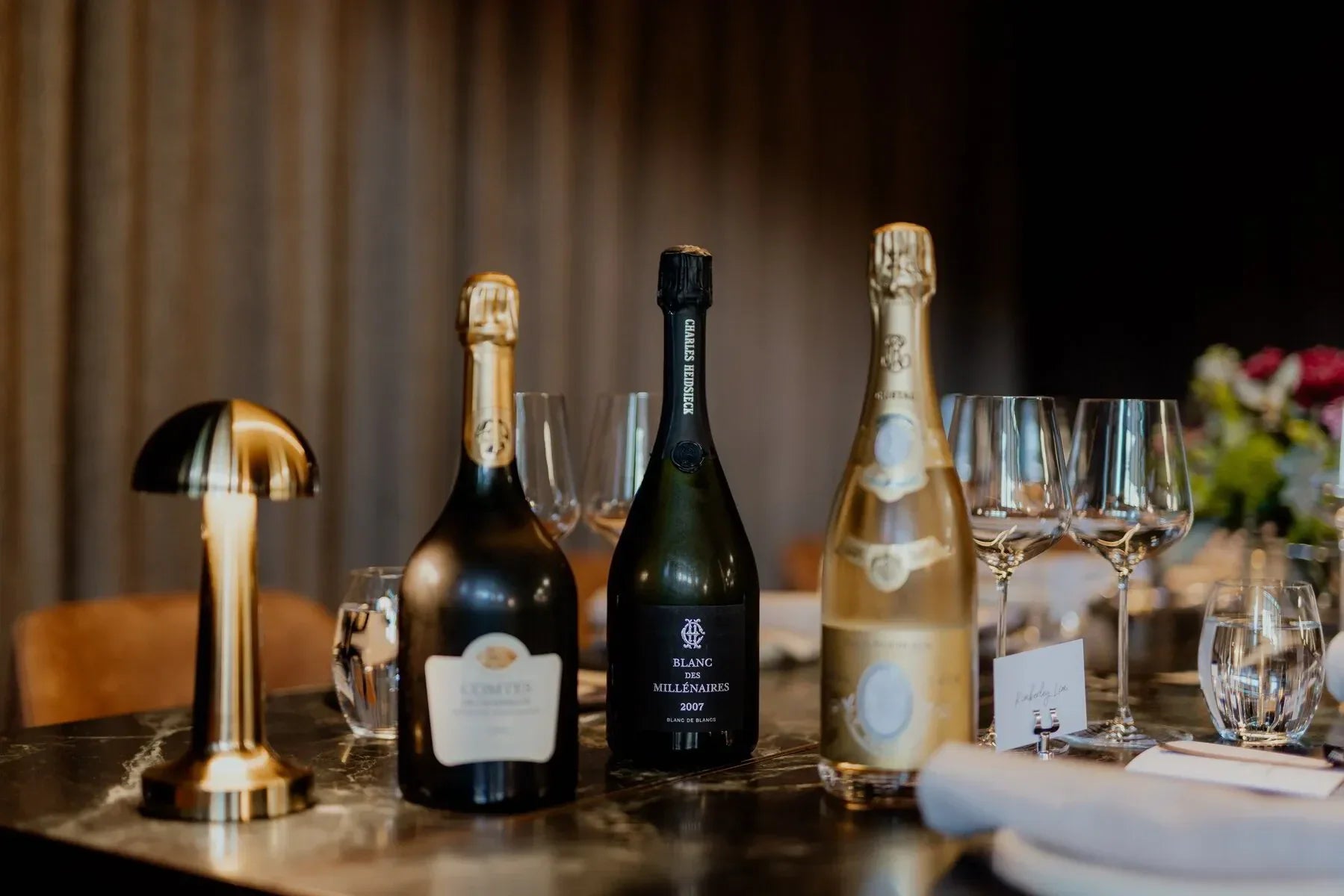 Three champagne bottles on elegant restaurant table with wine glasses and gold lamp