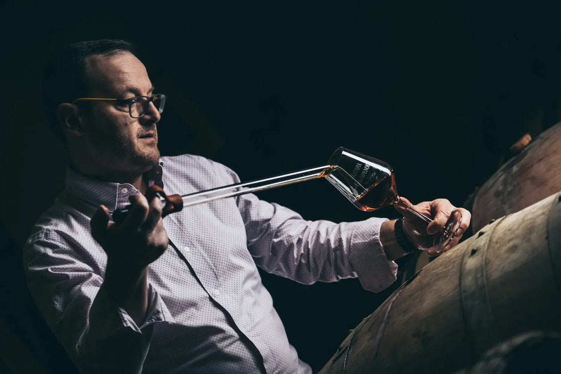 Man in glasses sampling whiskey from barrel with glass thief in dimly lit distillery.