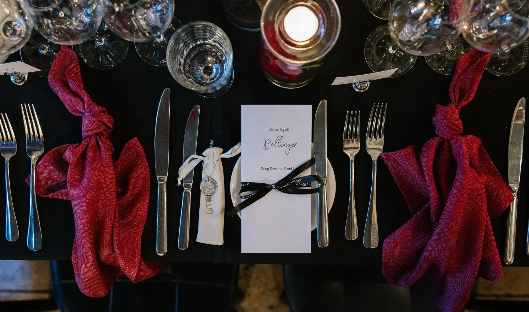 Elegant dinner table setting with red napkins, wine glasses, Bollinger menu, and candlelight.
