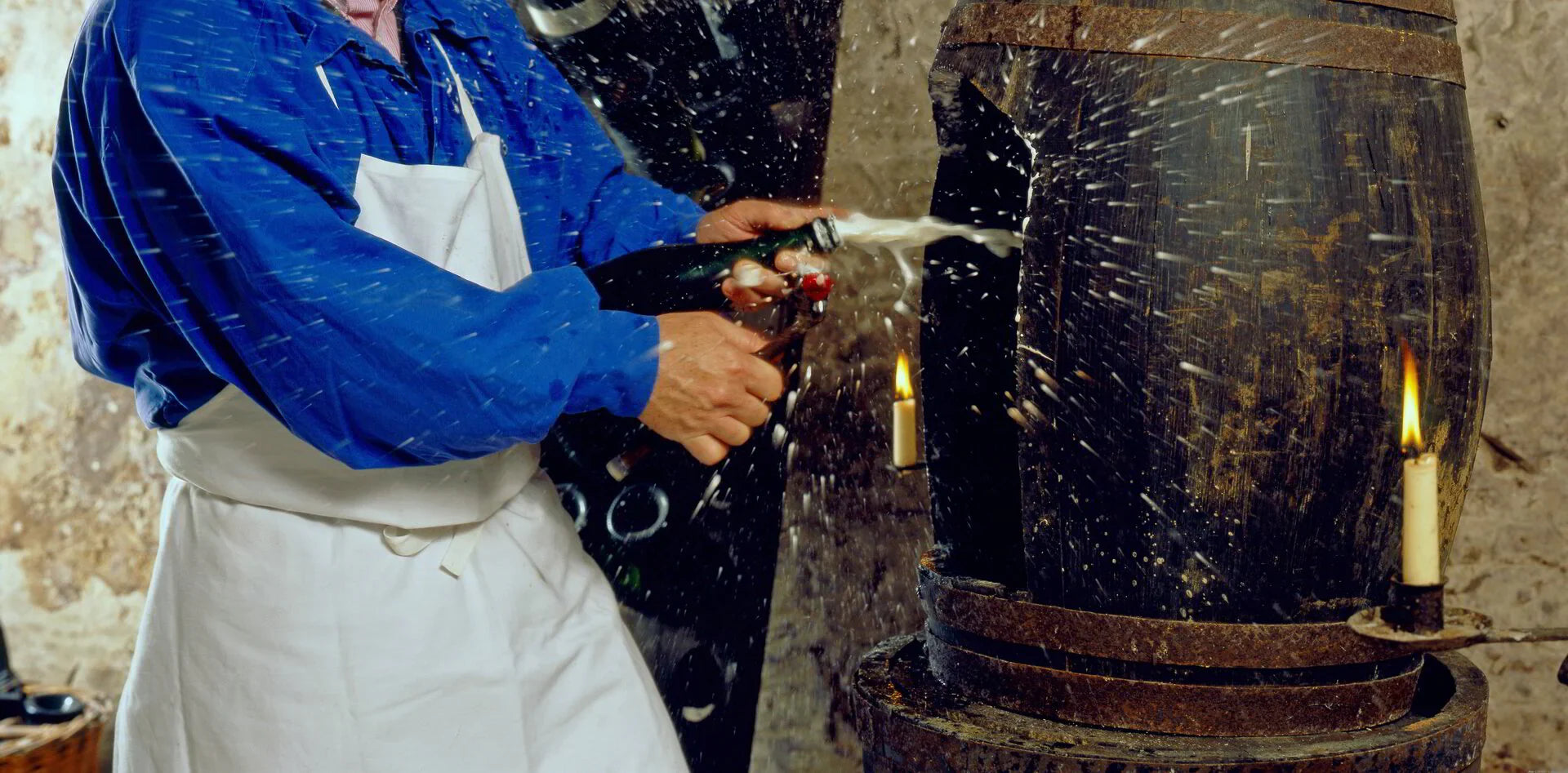 Worker in blue shirt and apron spraying champagne bottle in rustic cellar with barrel and candles