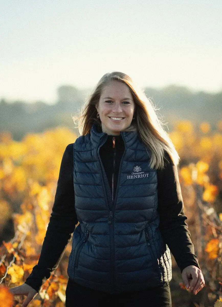 Smiling woman in Henriot vest standing in a sunlit vineyard in autumn.
