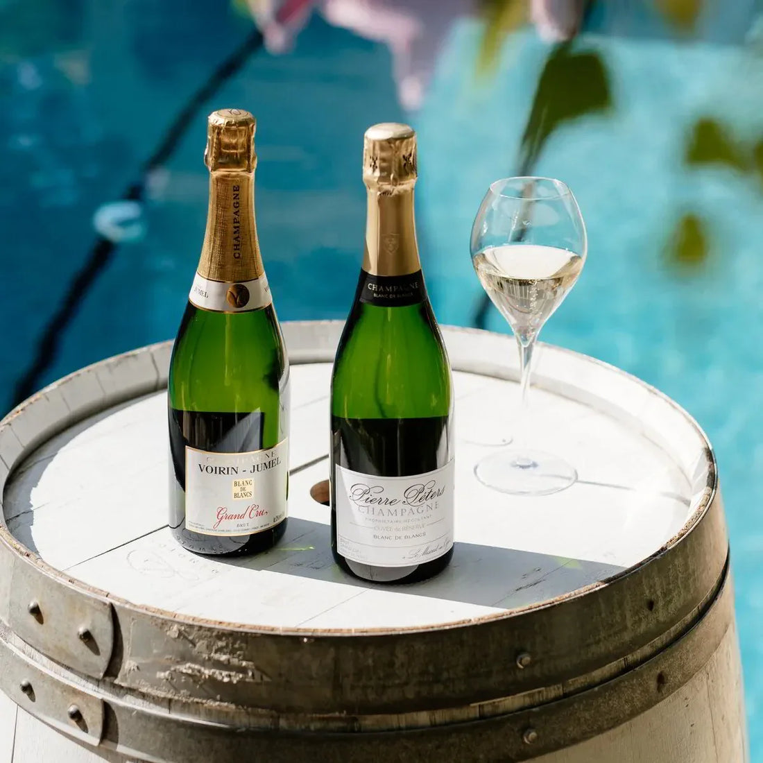 Two bottles of champagne and a wine glass on a barrel table by a pool