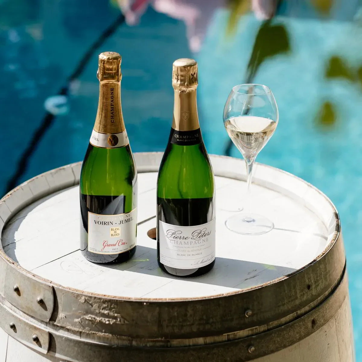 Two bottles of champagne and a wine glass on a barrel table by a pool