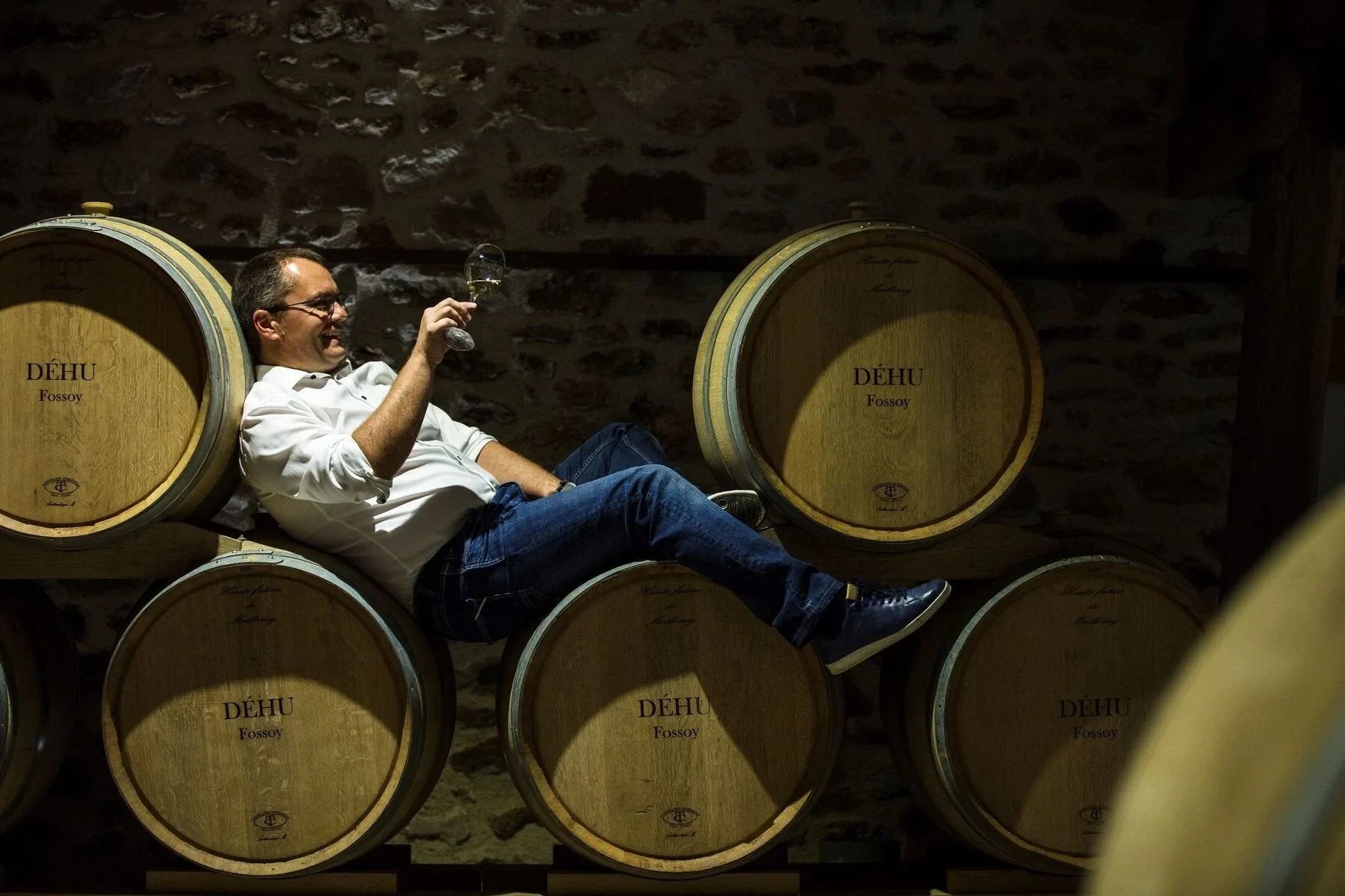 Man tasting white wine in a cellar, sitting on Déhu Fossoy oak barrels, stone wall backdrop