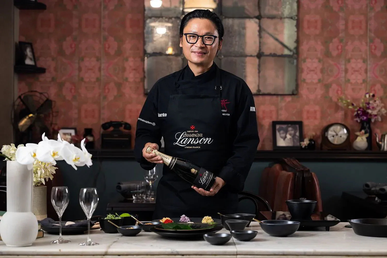 Chef in black apron holding Lanson champagne bottle in elegant kitchen with plated dishes