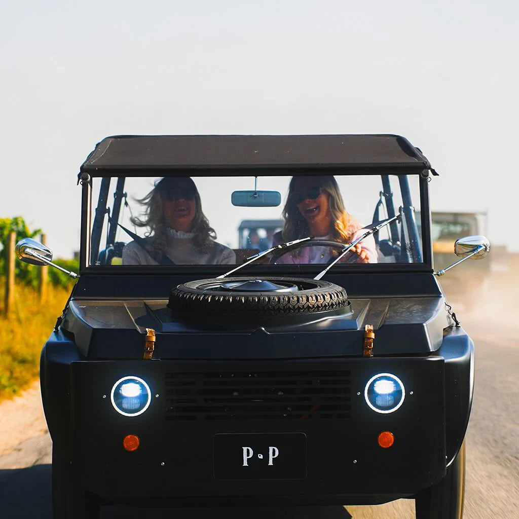 Two women driving a black vintage jeep with P P plate on a sunny vineyard road