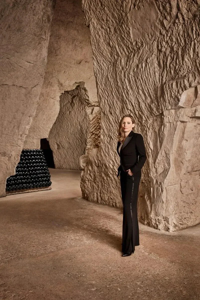Woman in black suit standing in stone wine cellar with champagne bottles stacked in background