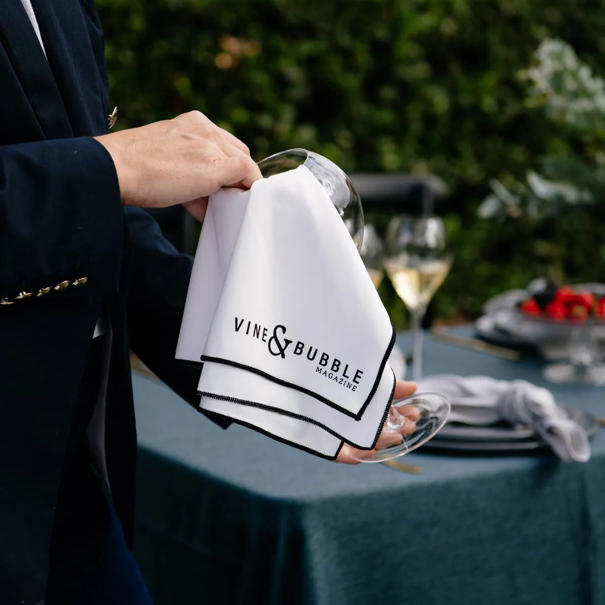 Polishing a champagne glass with a Vine & Bubble wine cloth at an outdoor table setting.
