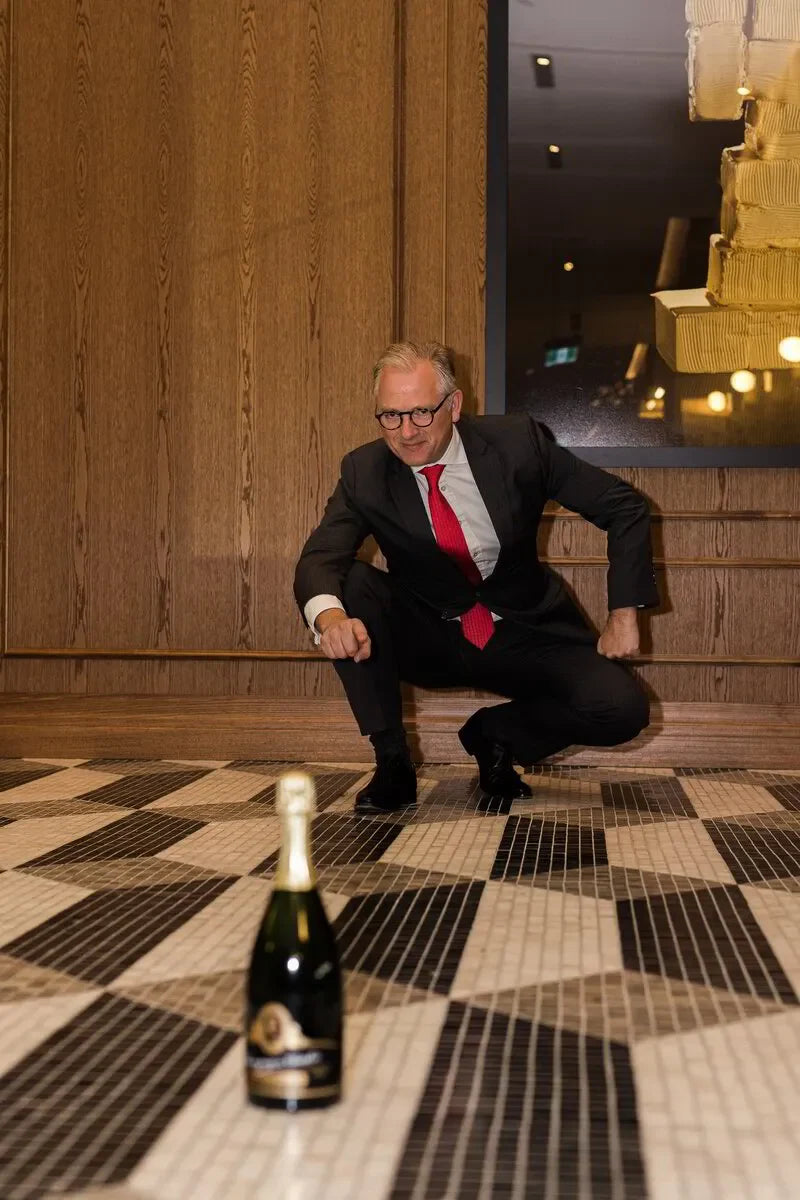 Man in suit with red tie crouching indoors on checkered floor near champagne bottle