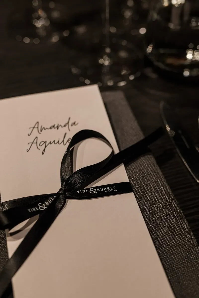 Elegant place card with handwritten name, black ribbon marked 'Vine & Bubble Magazine' on dark table