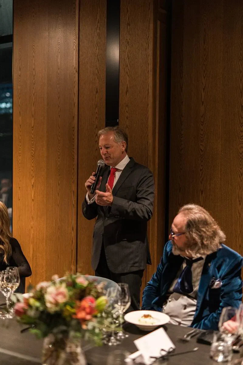 Man in suit giving speech at formal dinner event with flower centerpiece and wood panel backdrop