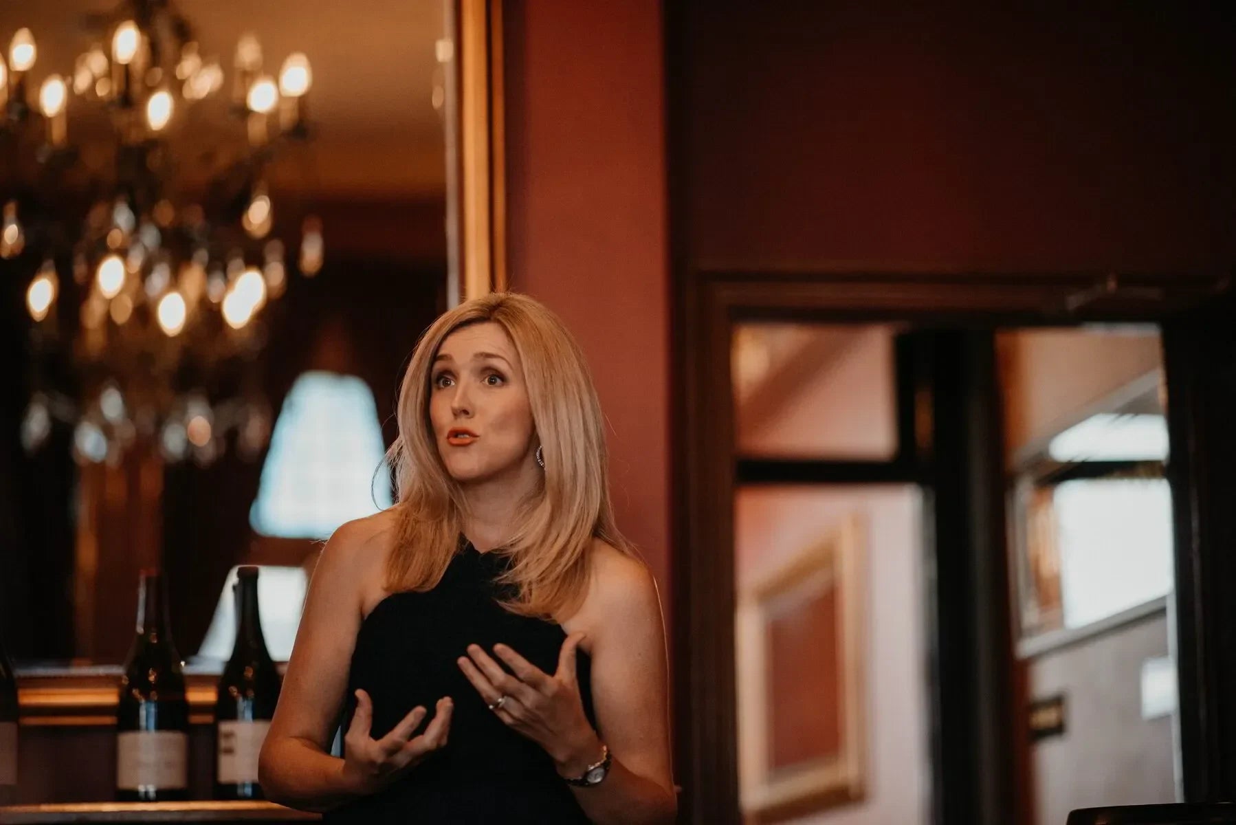 Blonde woman speaking indoors with wine bottles and chandelier, elegant event setting