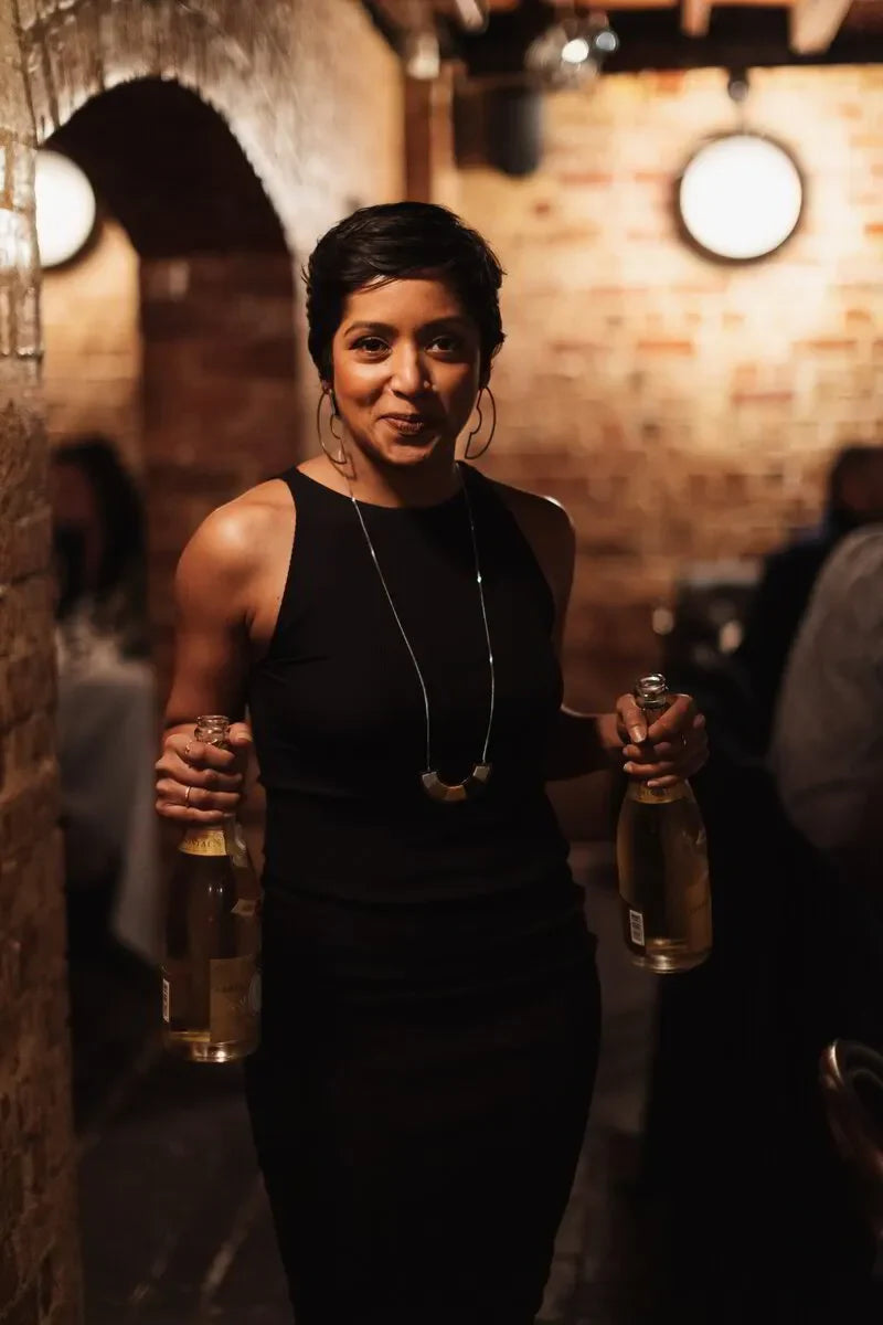 Woman in black dress holding two champagne bottles in a cozy brick restaurant setting