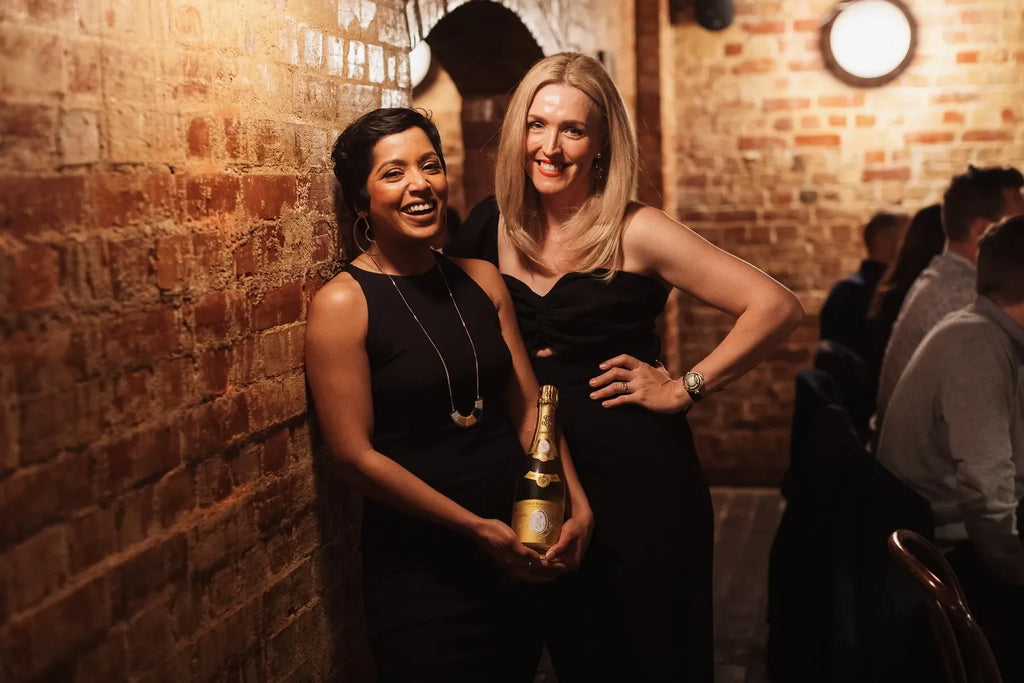 Two women in black dresses posing with champagne in a cozy brick-walled restaurant