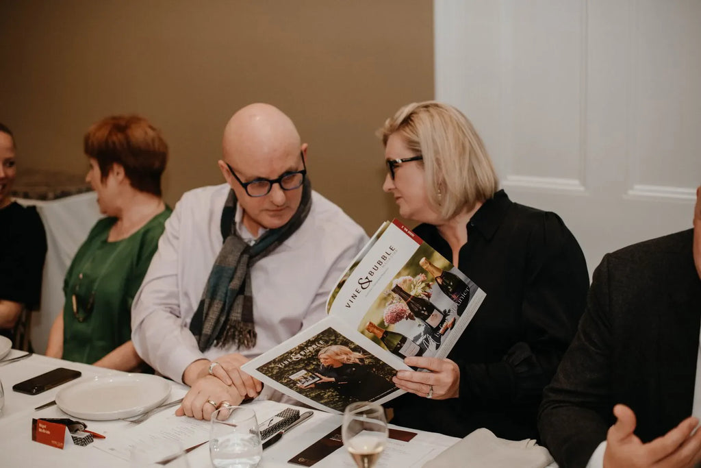 Man and woman at dinner table reading Vine & Bubble wine magazine, business event setting
