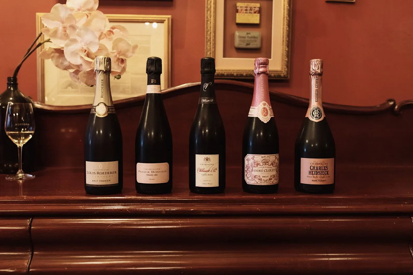 Five champagne bottles on wooden piano with framed art and glass of white wine