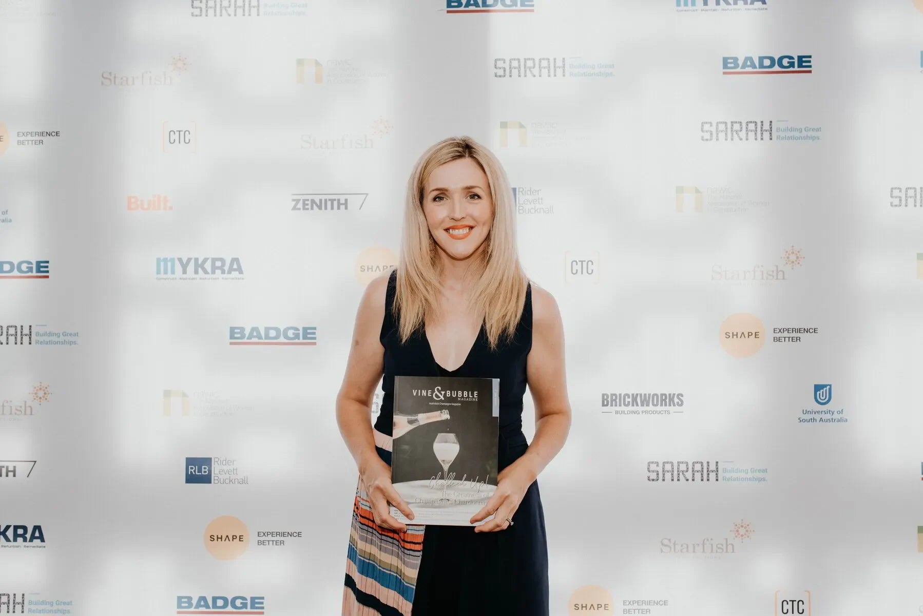 Smiling woman holding Vine & Bubble magazine at awards event with branded backdrop