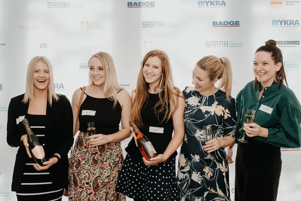 Five women smiling at an event, holding champagne, in front of sponsor backdrop