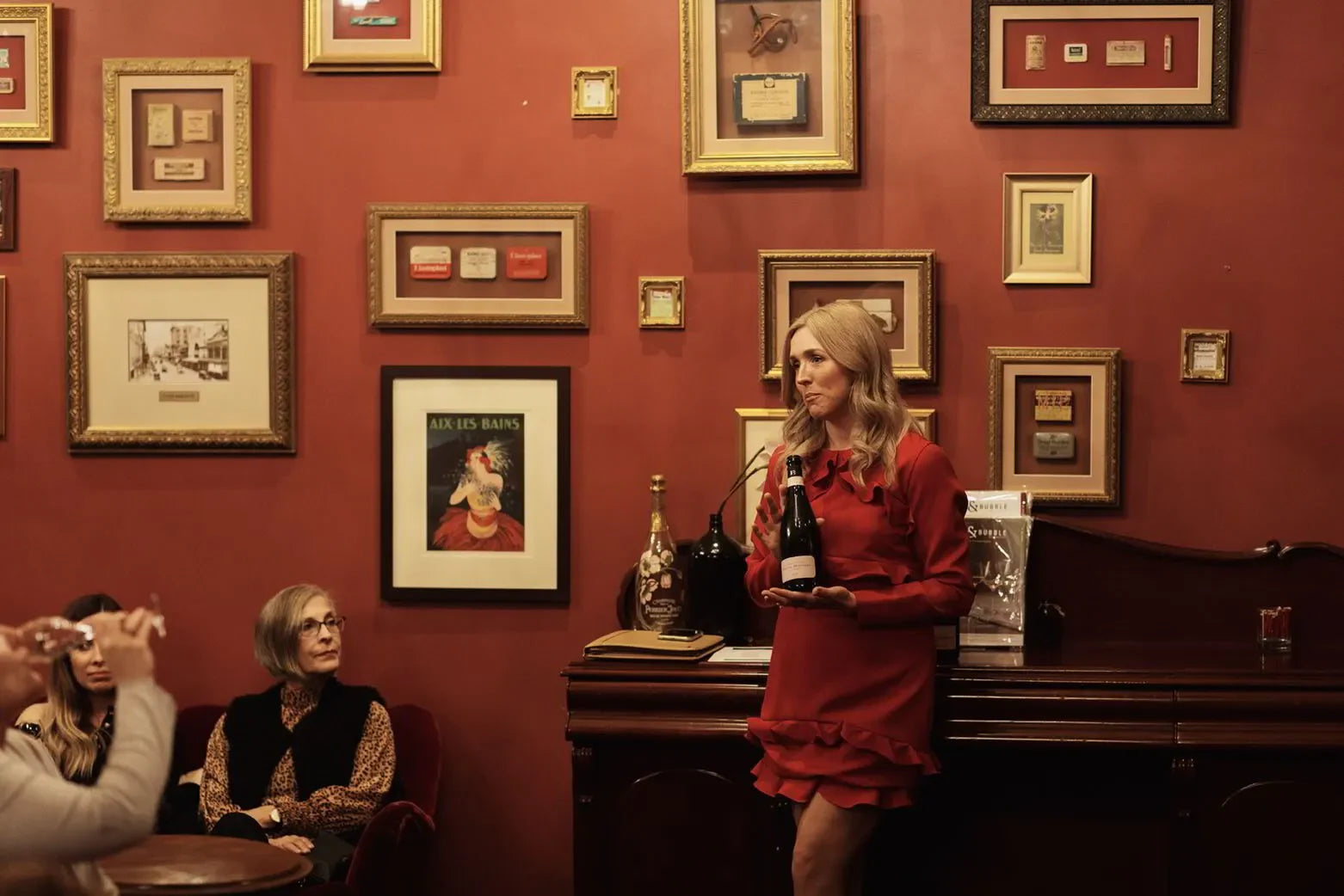 Woman in red dress holding wine bottle at vintage bar with framed art on red walls