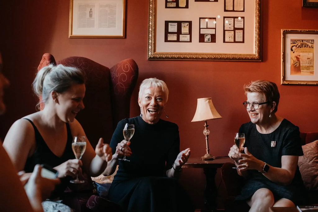 Three women laughing and holding glasses of champagne in a warmly lit lounge with framed art
