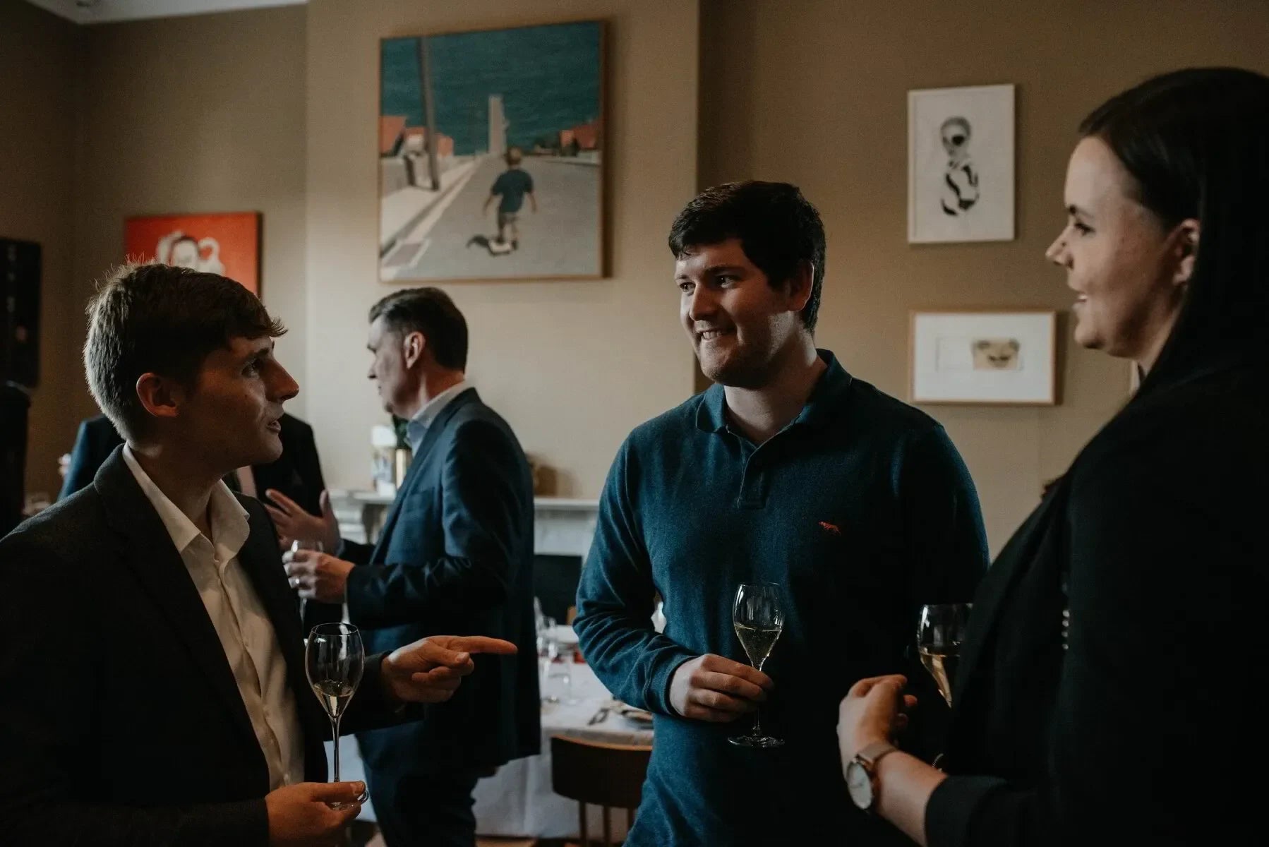 People networking at an indoor social event, holding wine glasses and talking in a gallery-style room