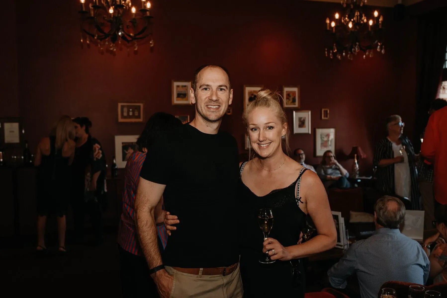 Smiling couple at indoor wine event with chandeliers and framed art on maroon walls