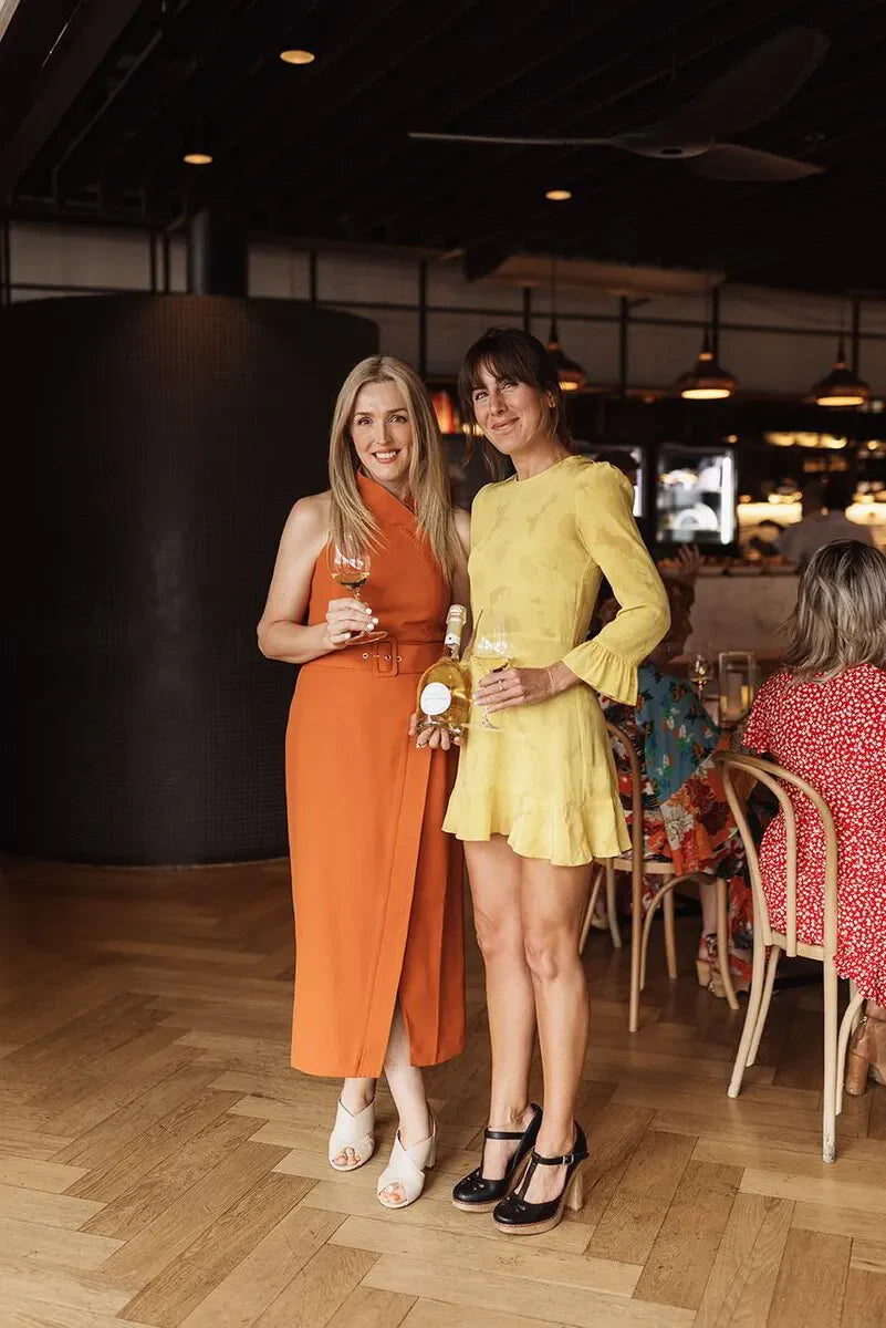 Two women dressed in bright dresses holding wine glasses and a wine bottle at an indoor event.