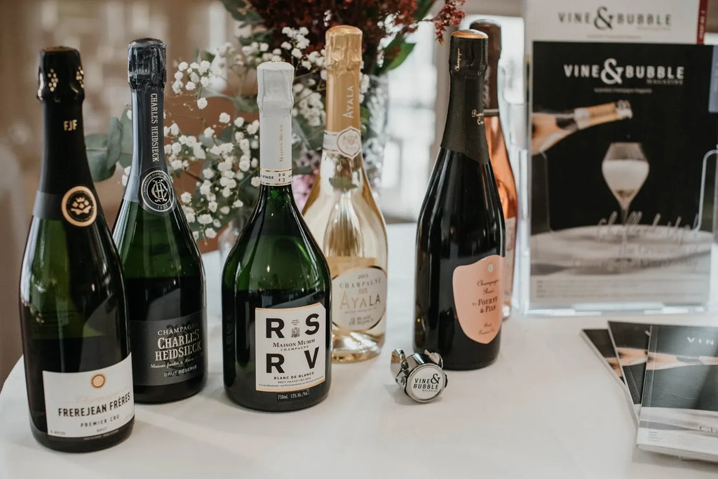 Assorted champagne bottles on table with Vine & Bubble brochures, floral decor in background