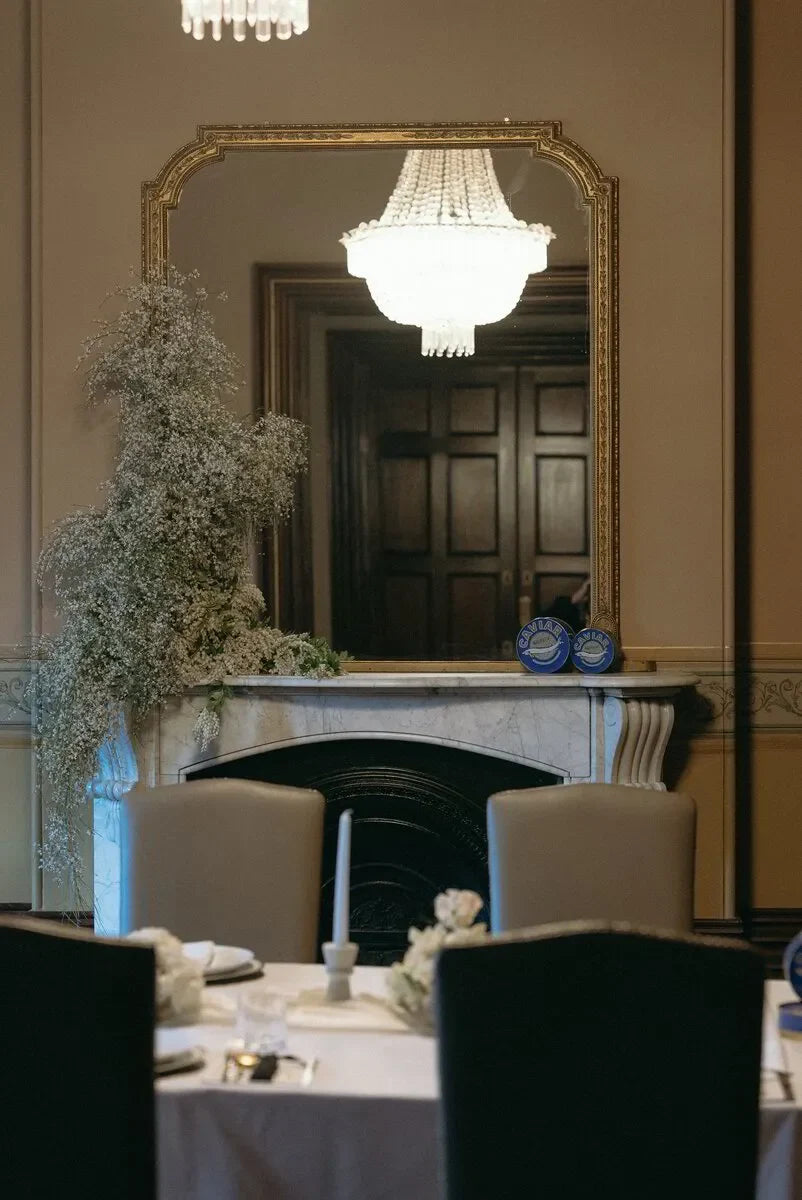 Elegant dining room with marble fireplace, large mirror, chandelier, and floral arrangement.