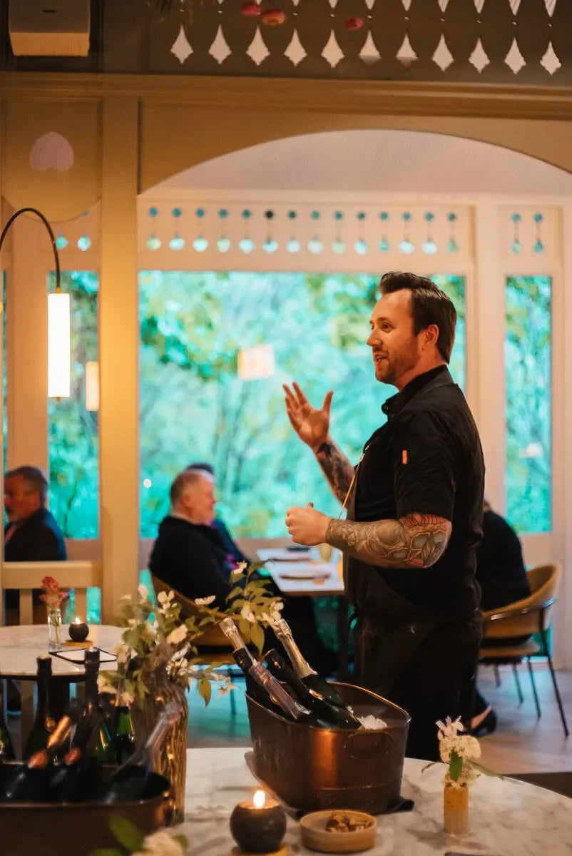 Tattooed man presenting wine in cozy restaurant with floral decor and natural light