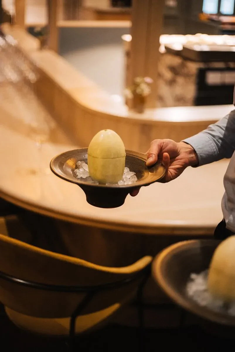 Hand holding a ceramic bowl with a yellow egg-shaped dessert on ice in a modern restaurant