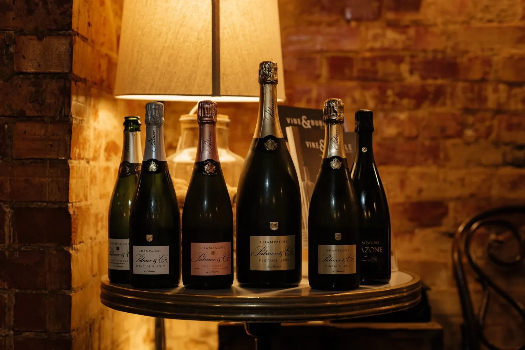 Assorted champagne bottles on a round table with warm brick wall and lamp background