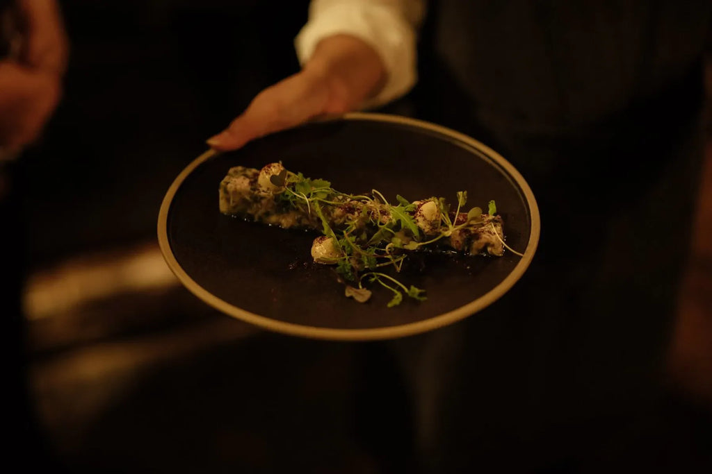 Hand holding a black plate with gourmet appetizer topped with microgreens in dim lighting