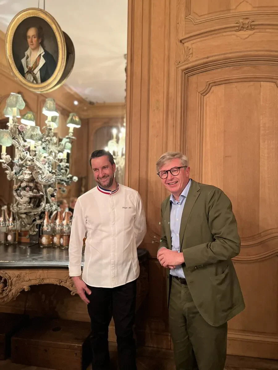 Chef in white coat and man in suit posing in elegant room with wood paneling and ornate decor