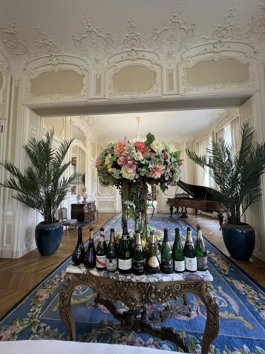Elegant parlor with ornate decor, marble table of champagne bottles, grand piano, floral centerpiece