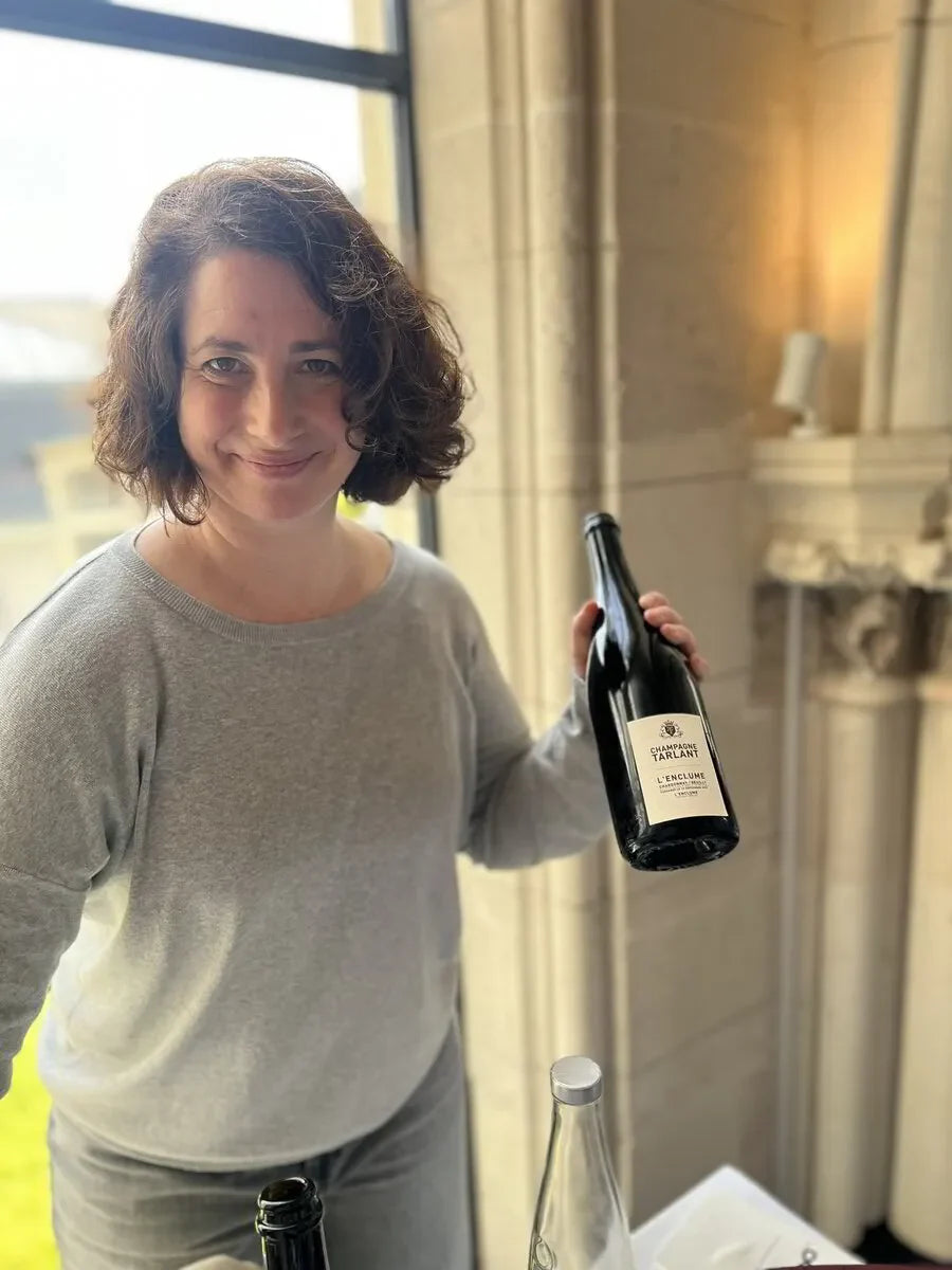Woman indoors holding a bottle of Champagne Tarlant L'Enclume, smiling by window.