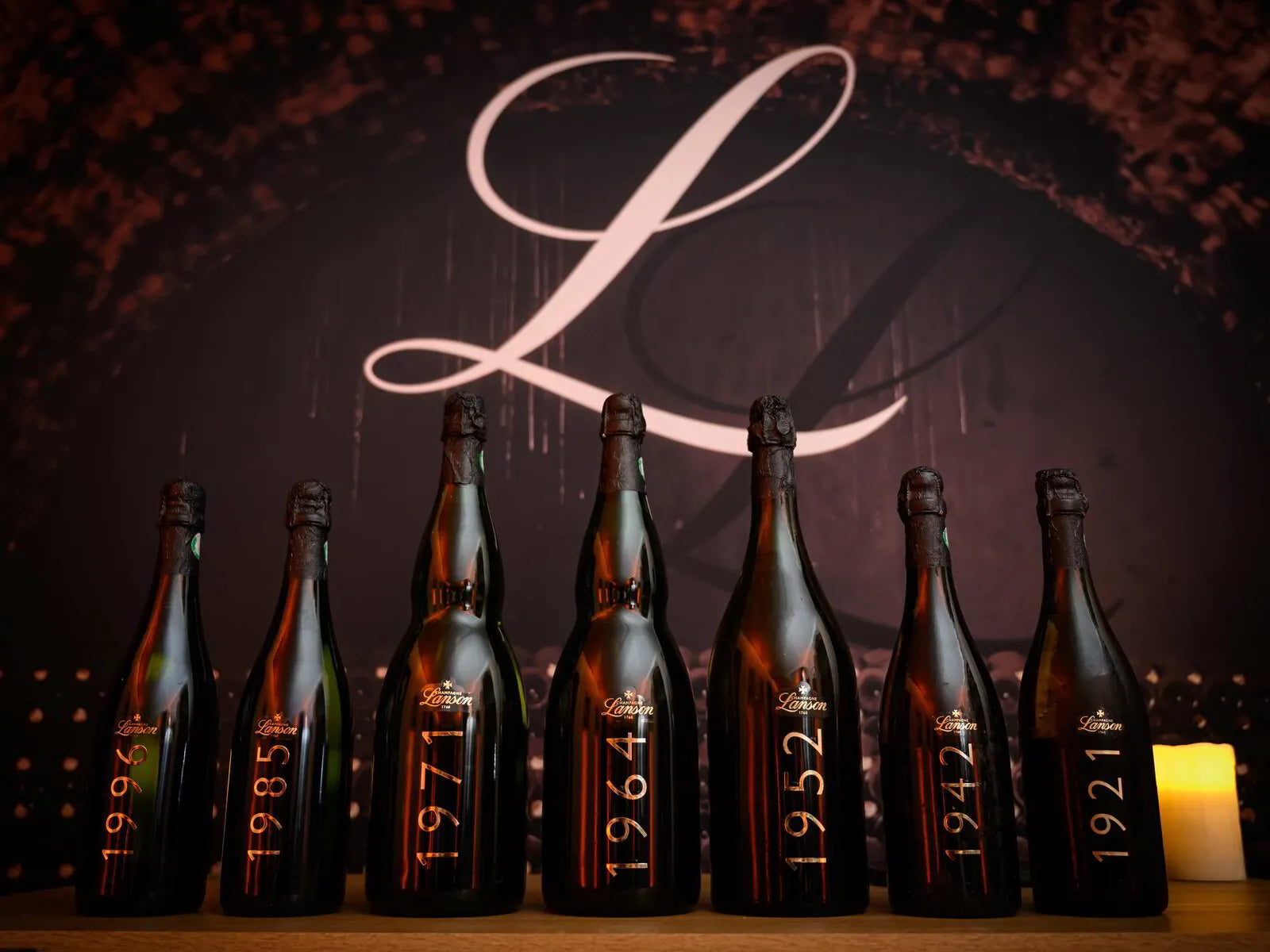 Seven vintage dark glass bottles with years from 1921 to 1999 displayed in a row with a large scripted L in the background