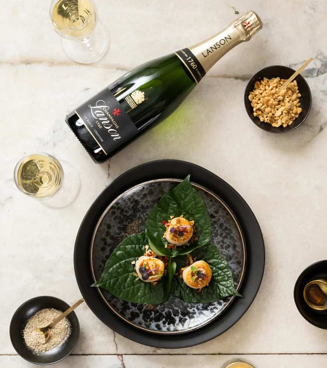 Bottle of Lanson champagne with three hors d’oeuvres on green leaves and two glasses of champagne on marble surface