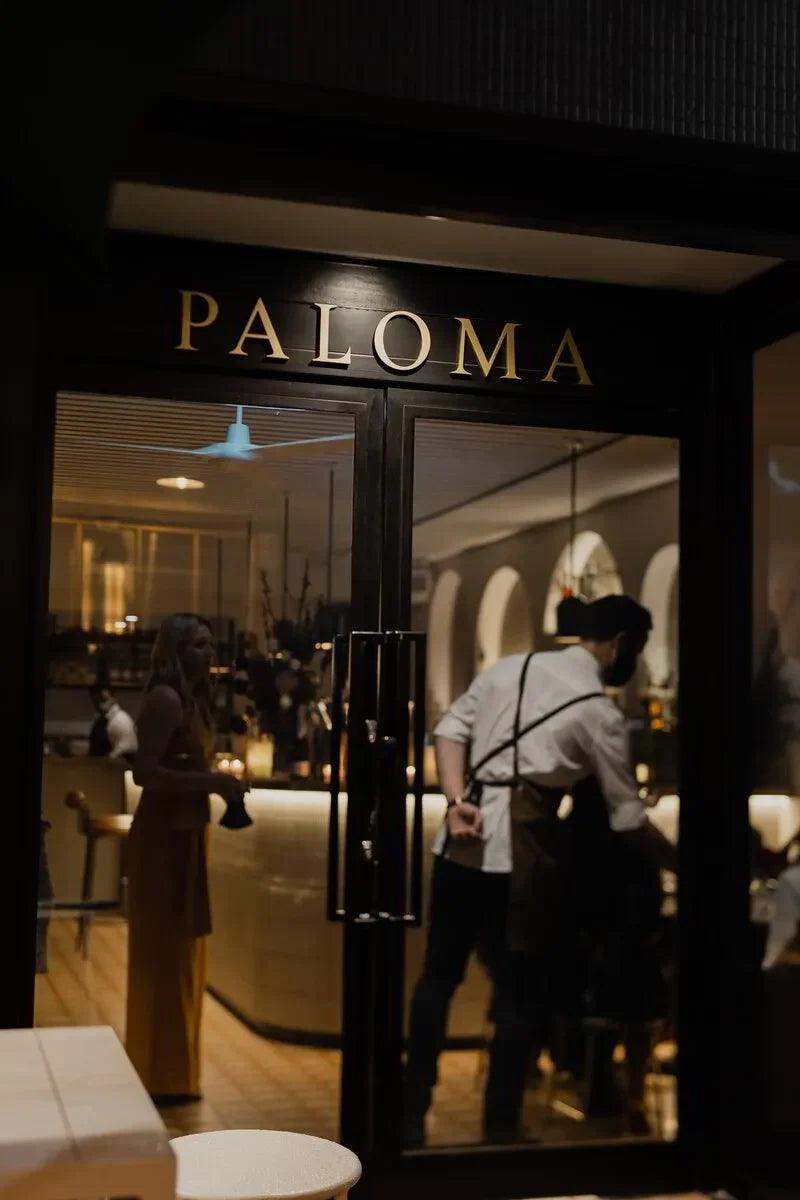 Entrance of Paloma restaurant with glass doors, visible staff and patrons inside, warm lighting