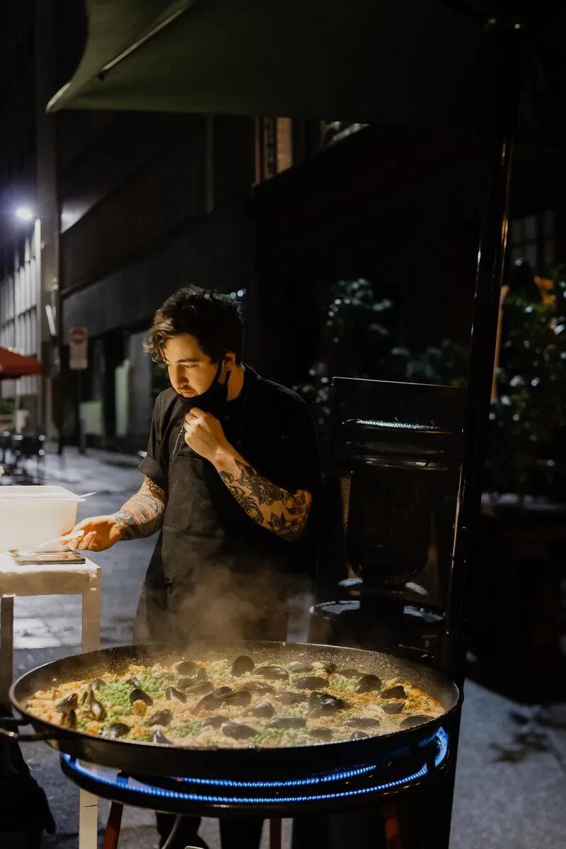 Tattooed chef cooking large seafood paella outdoors at night street market