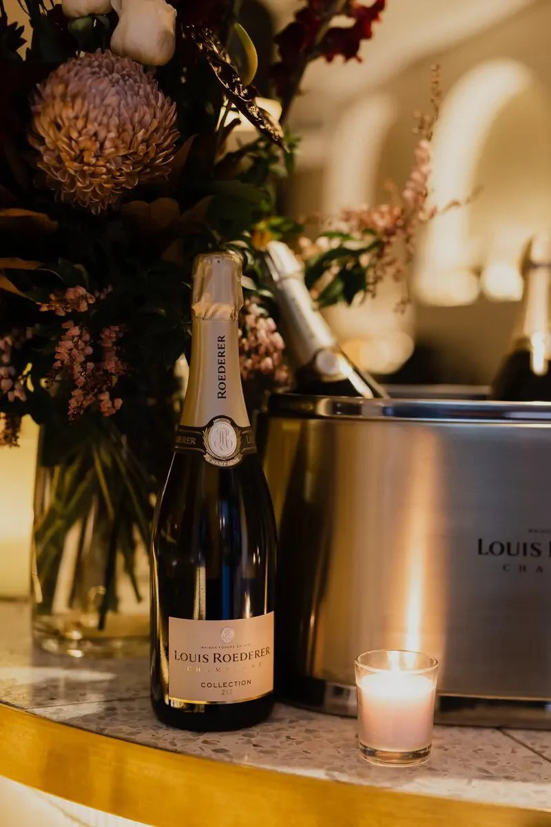 Louis Roederer Collection 242 champagne bottle beside flowers and a lit candle on a marble counter