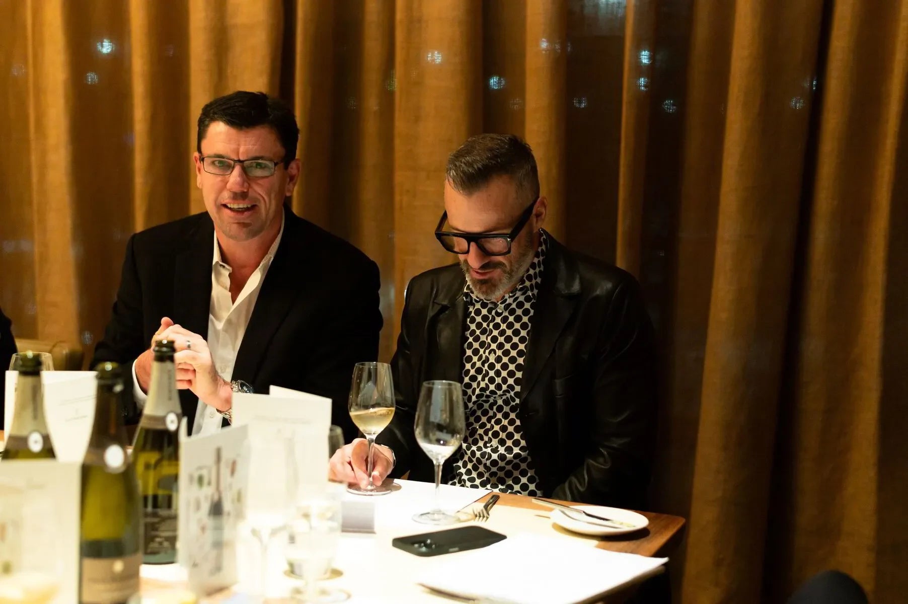Two men in suits at a restaurant table with wine glasses and bottles, brown curtain backdrop