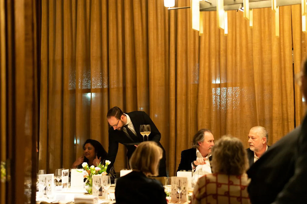 Fine dining event with guests seated at a table, server in suit holding wine glasses