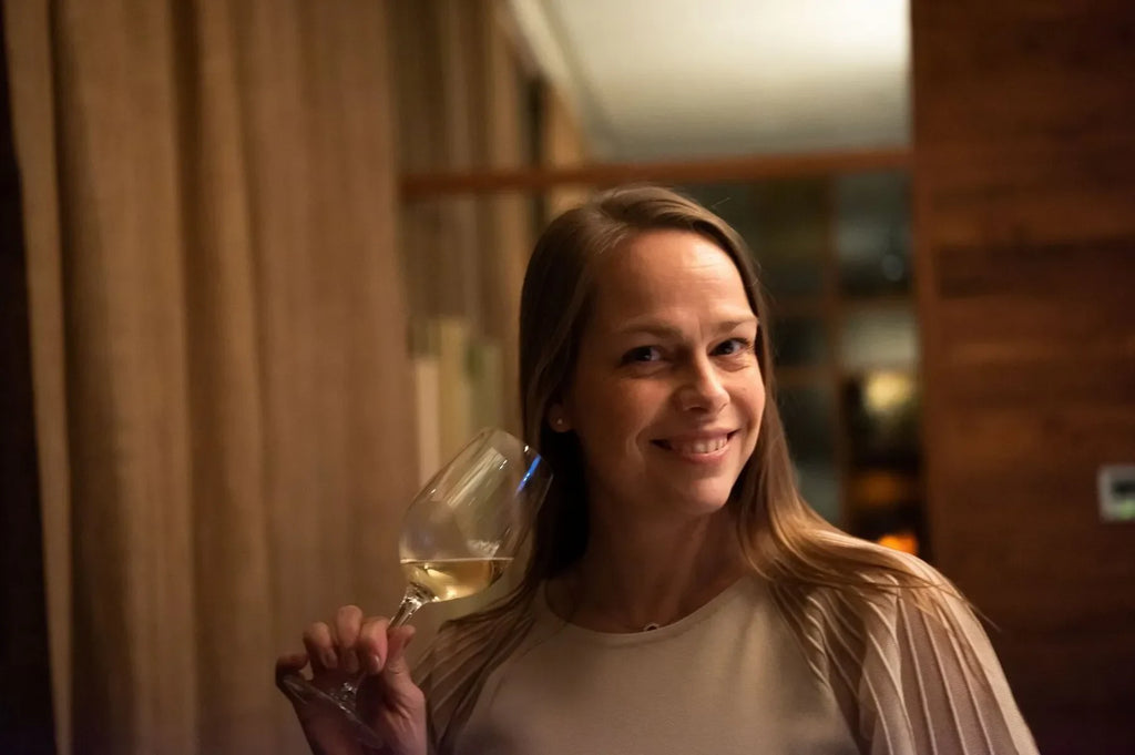 Woman smiling and holding a glass of white wine indoors with warm lighting.
