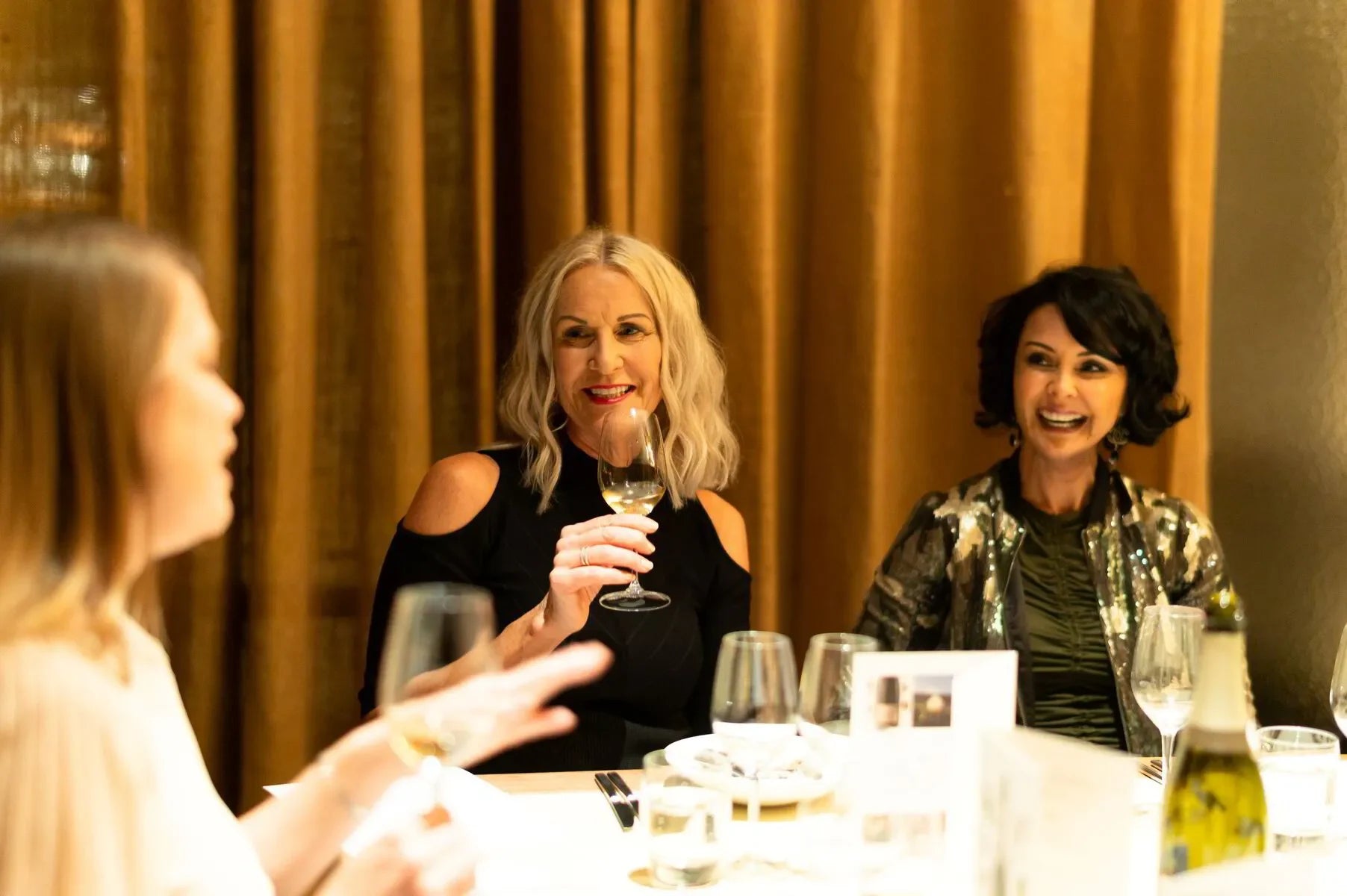 Three women enjoying wine at a table with gold curtains in an elegant dining setting