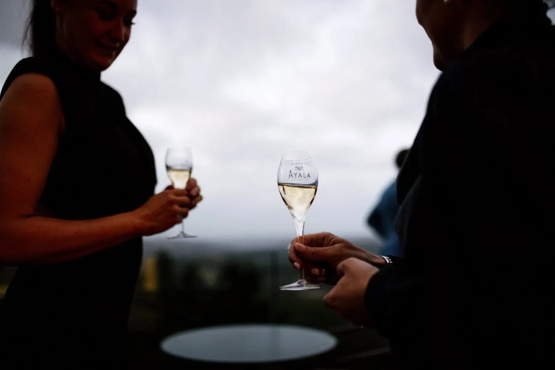 Two people holding Ayala champagne glasses outdoors at sunset with cloudy sky