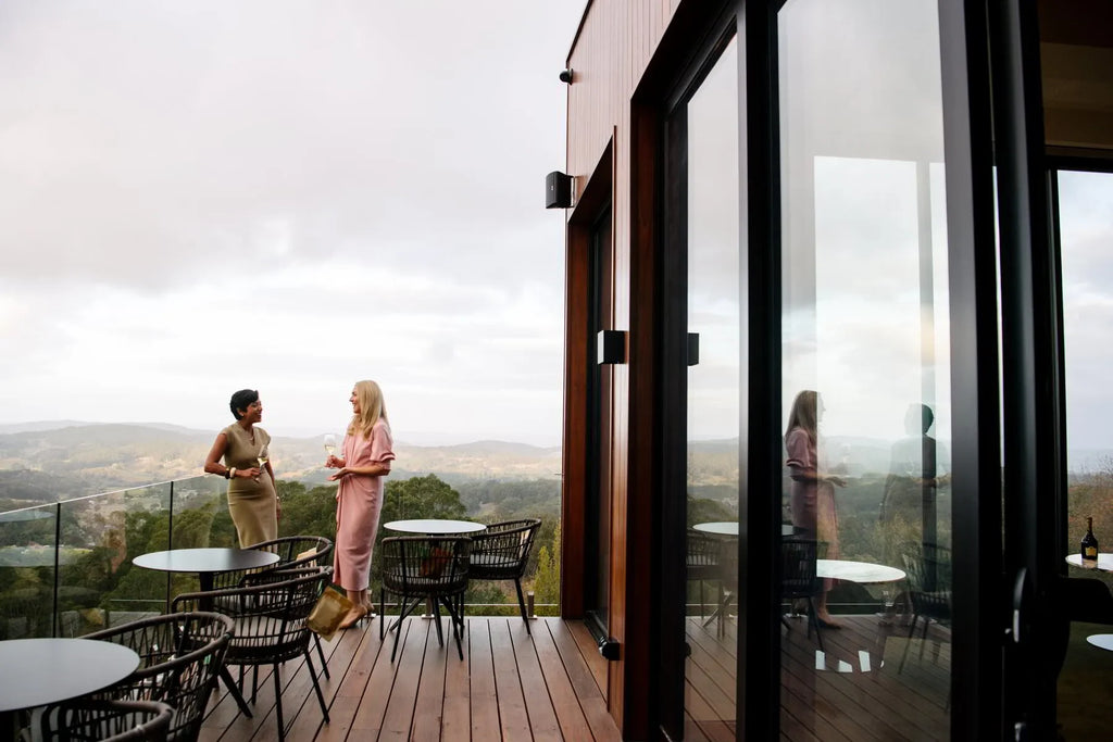 Two women on modern outdoor deck with wine, overlooking scenic hills and glass railing