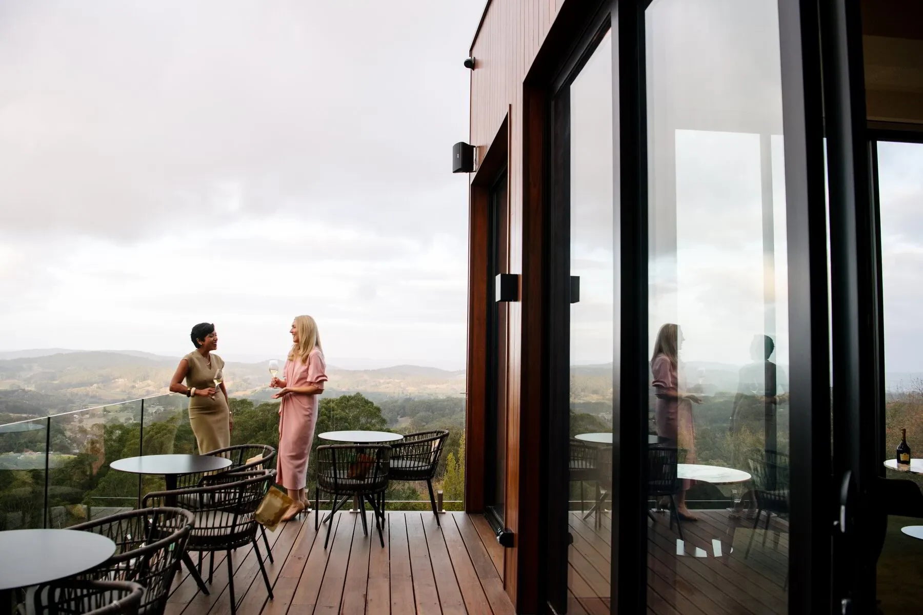 Two women on modern outdoor deck with wine, overlooking scenic hills and glass railing