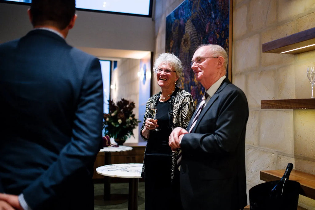 Elderly couple in formal attire socializing at a modern indoor event with wine and art decor.
